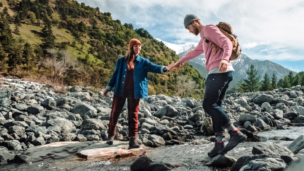 A woman helps a man cross a small river while hiking.