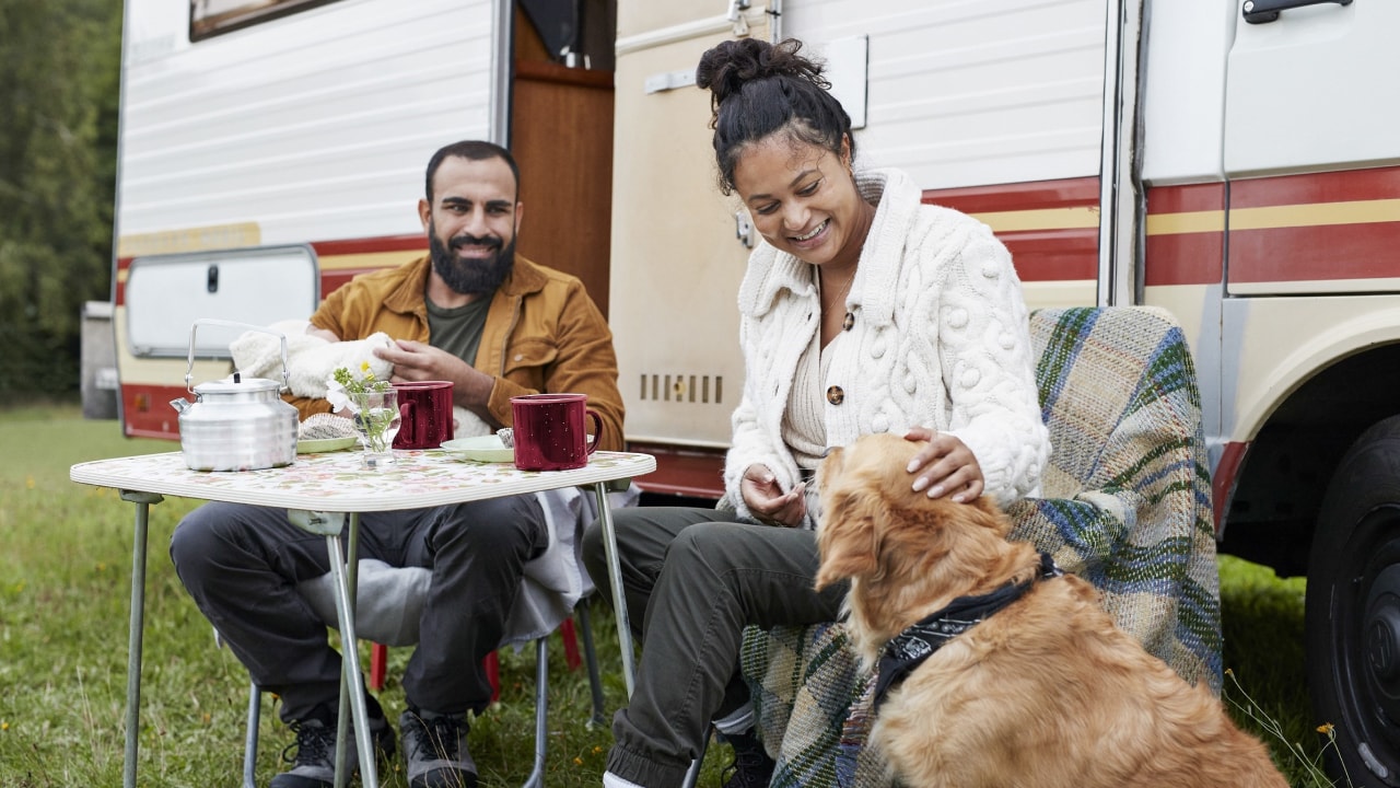 A husband and wife having lunch next to their caravan and dog.