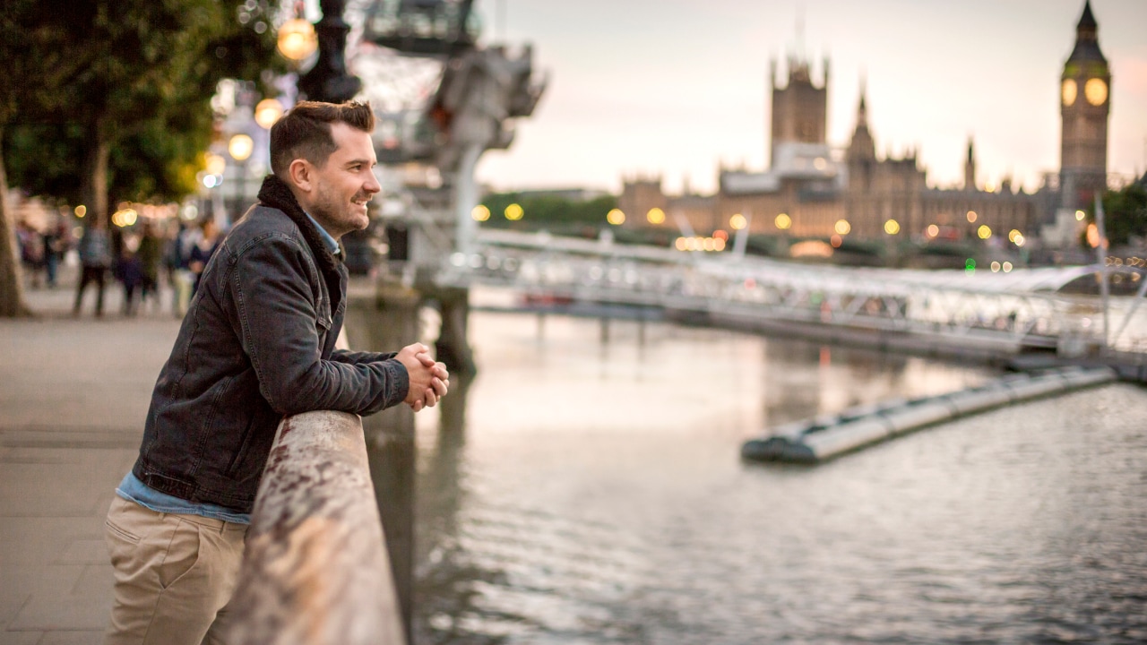 A man leaning over railing looking at a river with a city in the background.