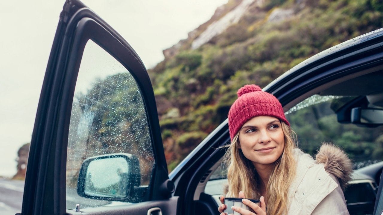 A woman wearing a beanie, holding a drink, while sitting in a car next to a mountain.