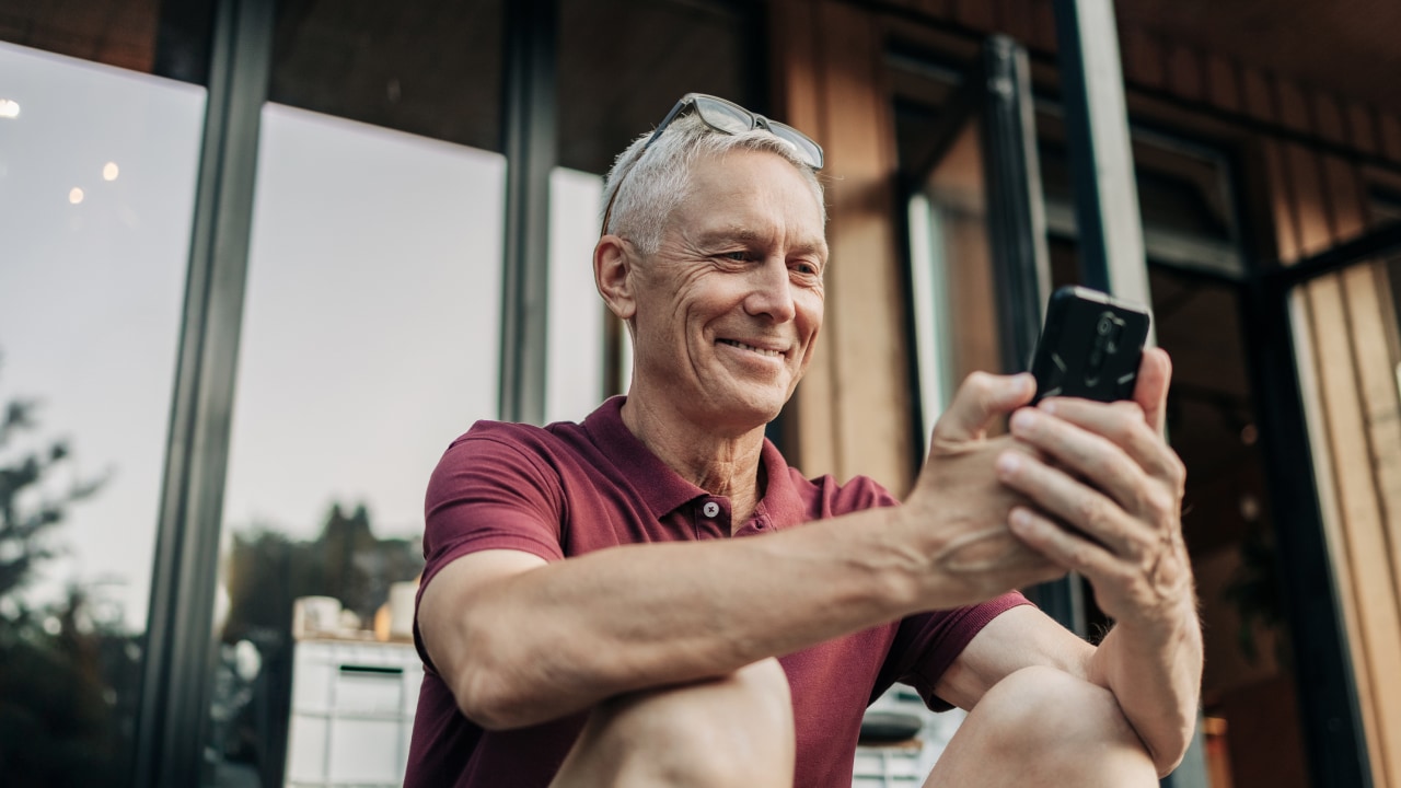 An elderly man sitting outside smiling while looking at his phone.