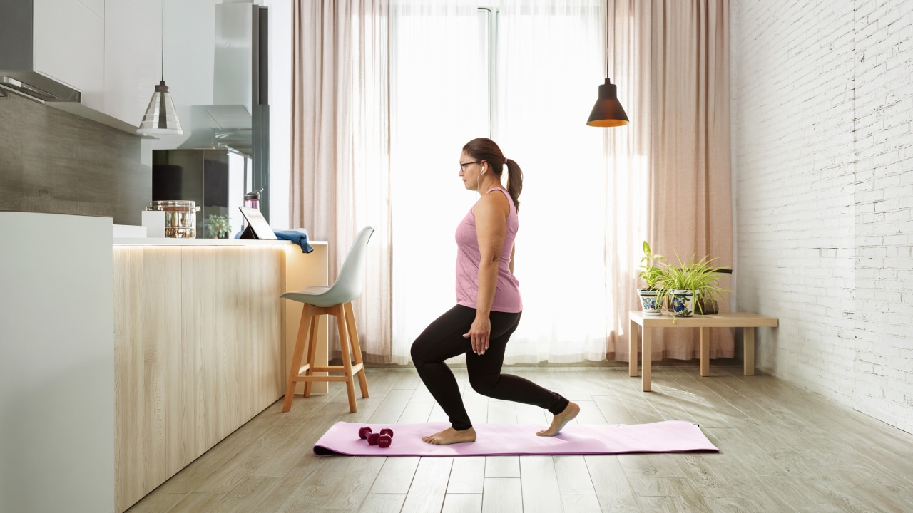 A woman exercises on a gym mat while watching a tablet on the kitchen bench.