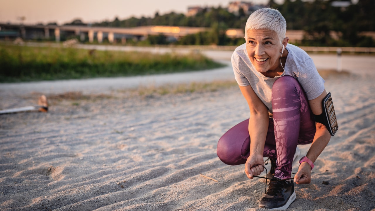 An elderly woman wearing running clothes tying her shoe on the beach.