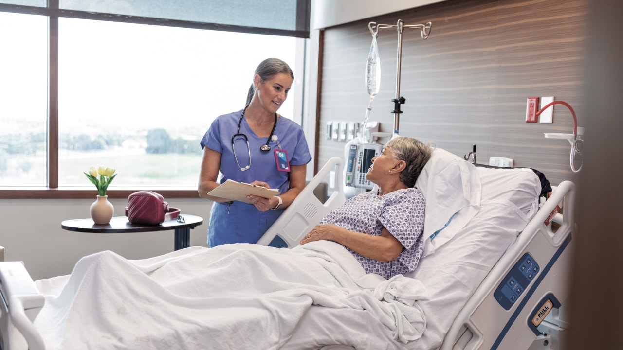 An elderly woman reclines at an angle in a hospital bed as she speaks to a medical specialist holding a clipboard.