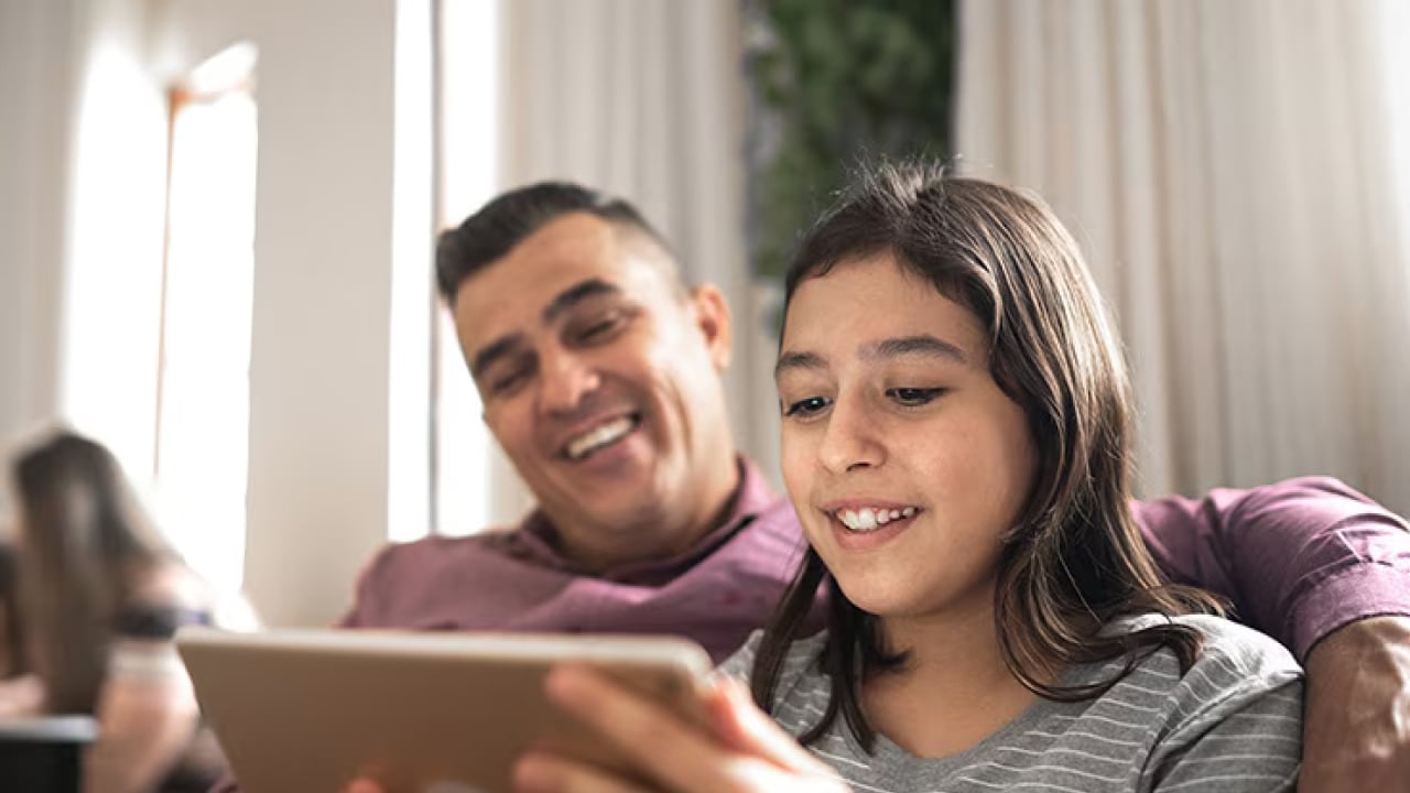 A girl smiles as she sits on a sofa and uses an app on her tablet with her father in the background. 