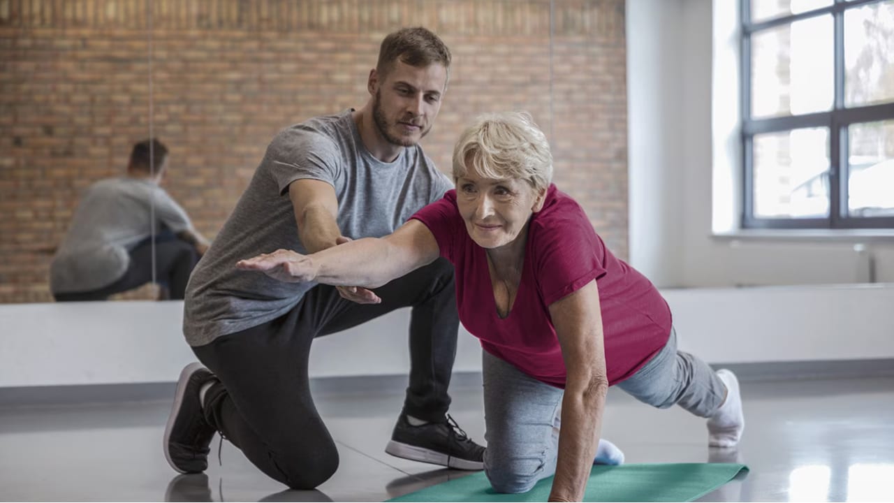 A senior woman does a yoga pose on one hand and one knee with the help of a male trainer.