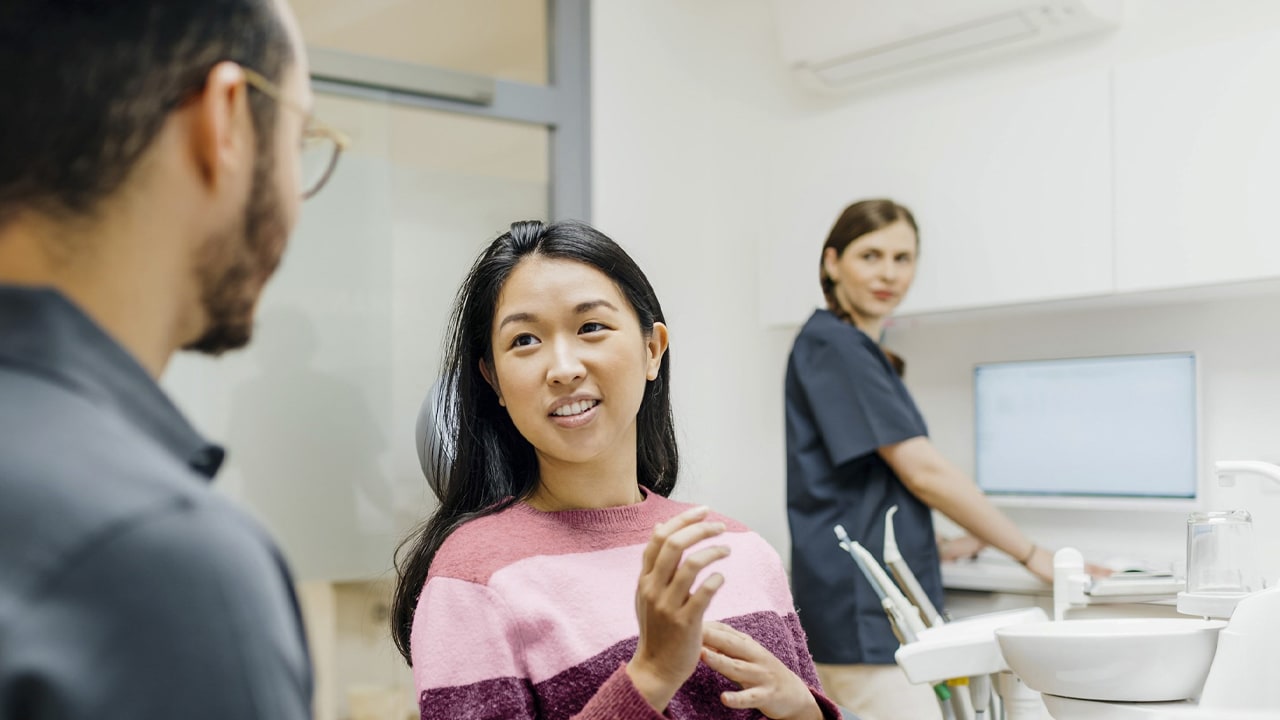 A woman sitting upright in a dental chair talks with a male dentist with an assistant behind.