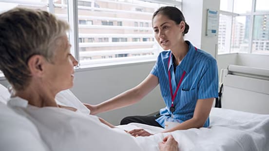 A female doctor sits on a hospital bed while talking to and comforting a female patient.