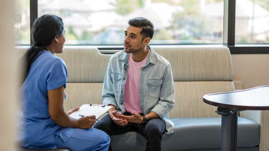 A patient sits on a couch talking to a medical professional holding a clipboard.
