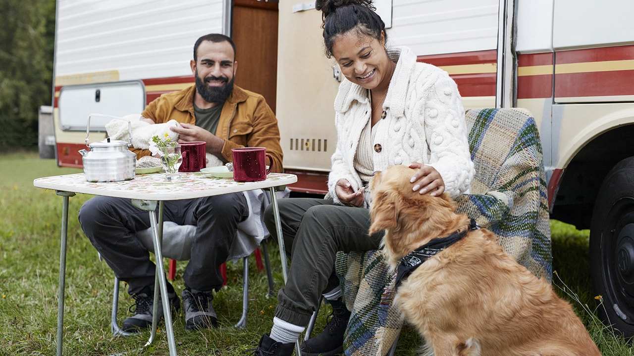 A man and woman having lunch outside their caravan with their dog next to them.