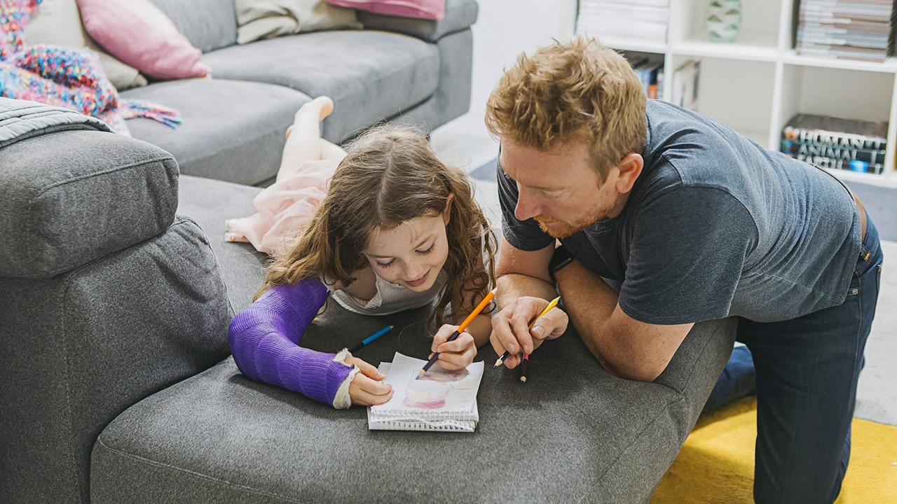 A father and daughter laying on the lounge doing homework.