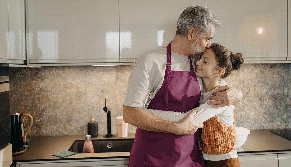 A father kissing his daughter on the forehead, standing in a kitchen.