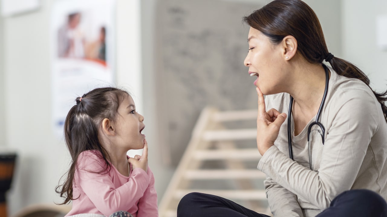 A woman wearing a stethoscope points to her chin while a child does the same next to her.
