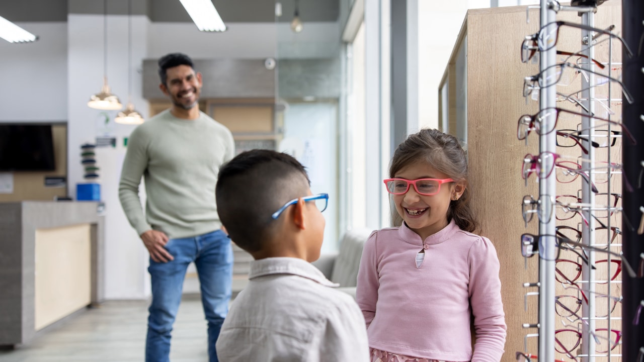 Two kids trying on glasses at an optometrist while their father smiles watching them in the background.