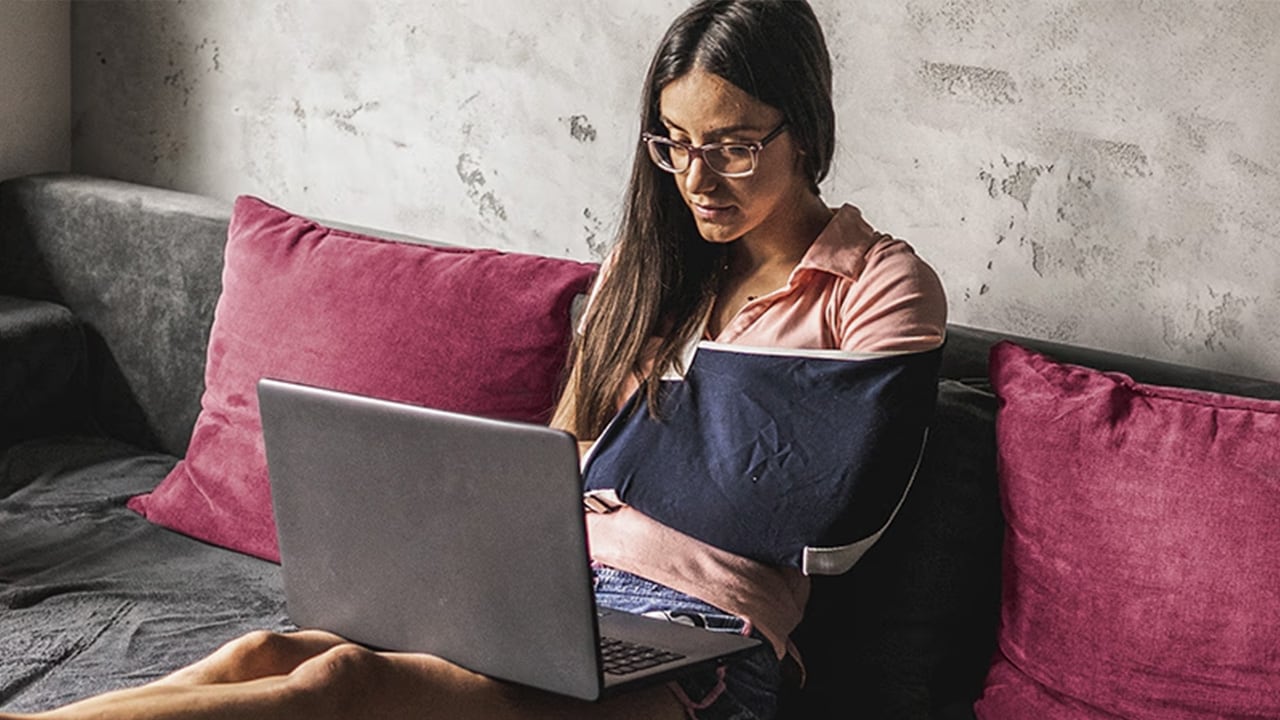 A lady wearing an arm sling while working on a laptop on her lounge.