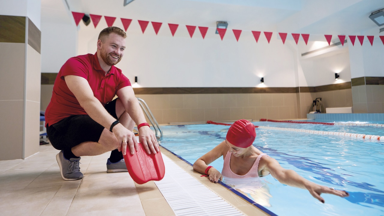 A lifeguard holding a floatation device crouching down next to a woman swimming in a pool.