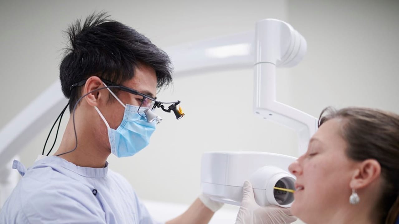 A male dentist sets up an x-ray machine while a female patient waits with her eyes closed.
