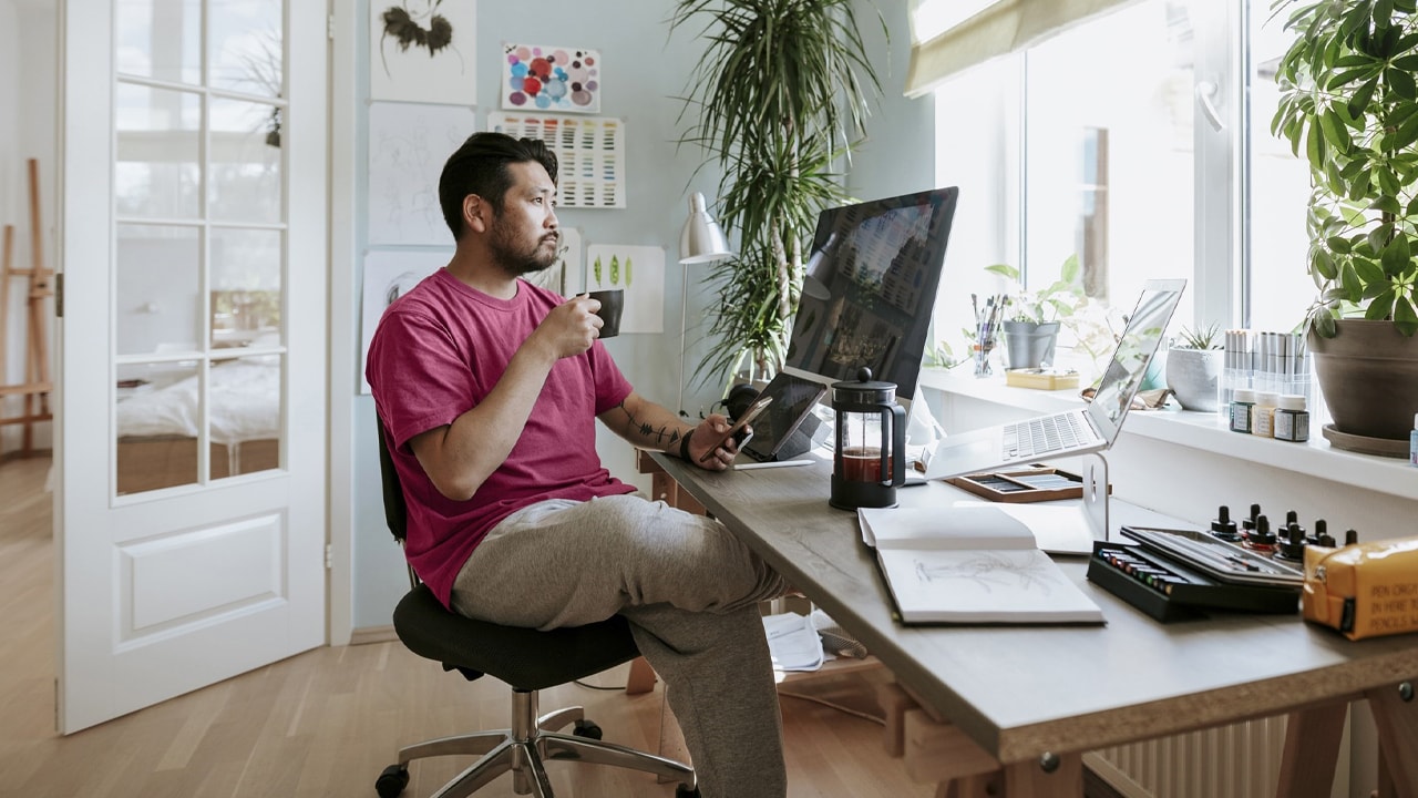 Man sitting at a desk in an office with a coffee and phone in each hand looking at a computer.