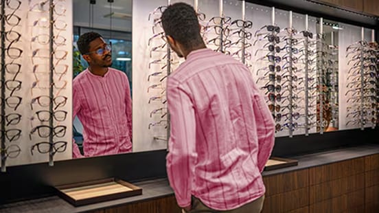 A man facing a wall of glasses looks at his reflection in a mirror while trying some on.