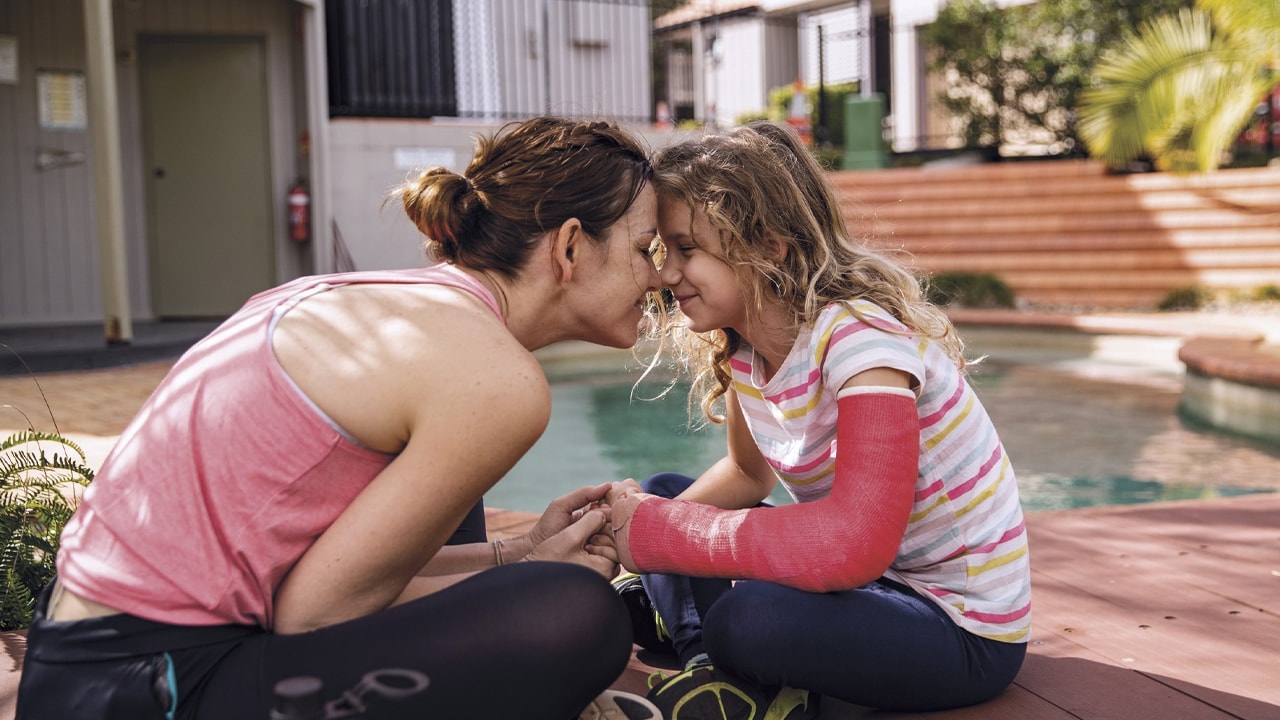 A mother and daughter sitting outside by a pool with their foreheads pressed together.