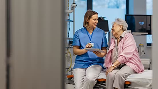 A female nurse holding a clipboard sits on a bed with an elderly patient in a hospital.