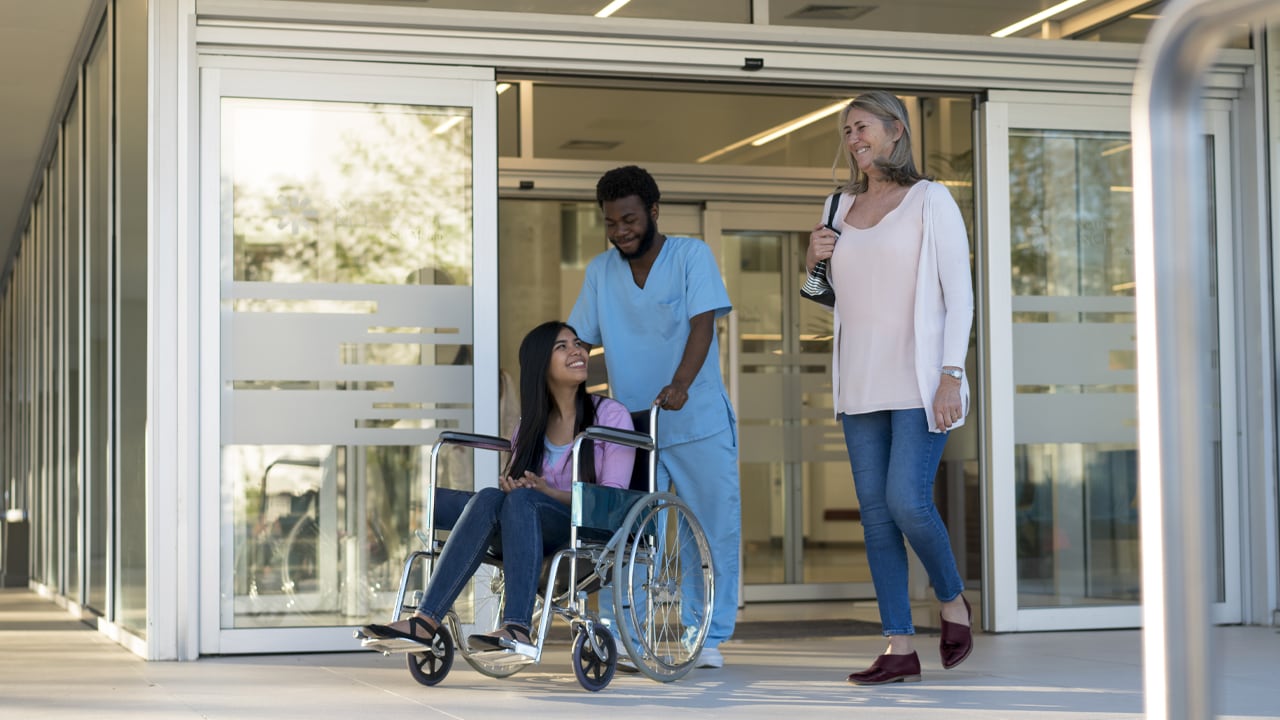 A nurse pushing a wheelchair with a young woman sitting in it while her mother walks next to her.