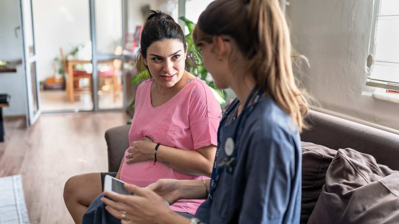 A photo of a pregnant woman sitting on her couch at an in home consultation with a nurse.