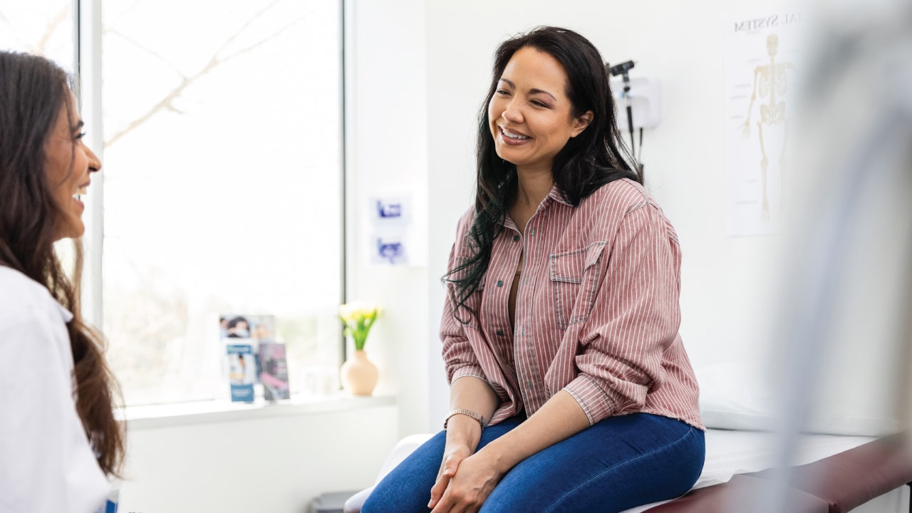 A woman is smiling, sitting on a hospital bed, speaking to a medical specialist.