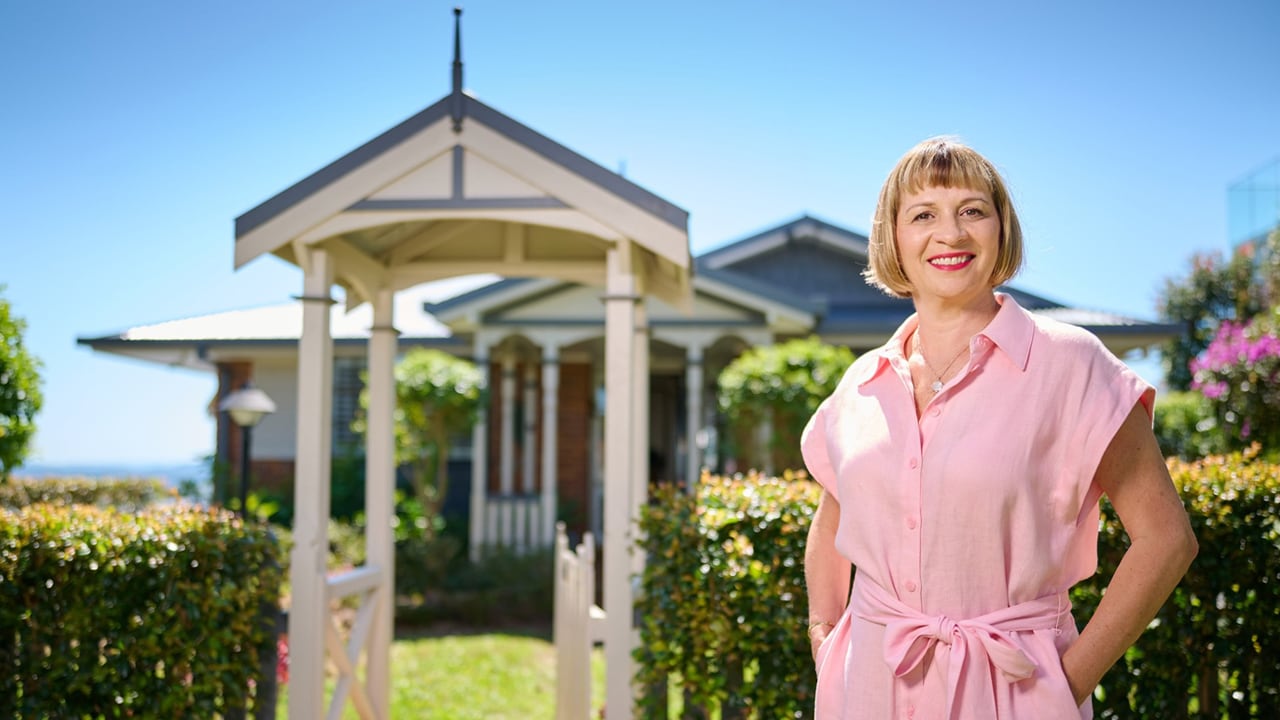 A woman smiling wearing a pink jumpsuit standing in front of her home on a sunny day.