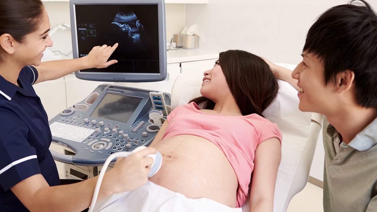 A pregnant woman lies back on a clinic bed with her partner standing next to her while a doctor or nurse performs an ultrasound and points to the screen.