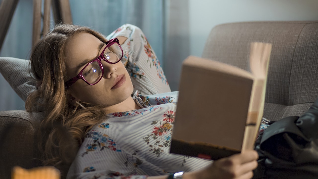 A young woman wearing glasses lying on the couch while reading a book.