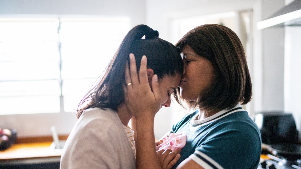 A woman kissing another woman on the forehead standing in a room.