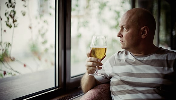 A bald man looking outside of a window while holding a drink